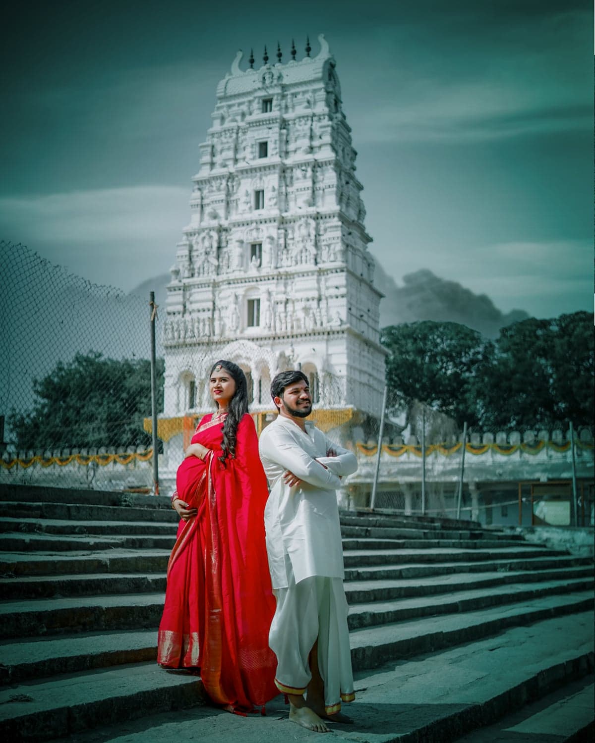 Maternity announcement photo of the couple in a red saree and white kurta before a heritage temple